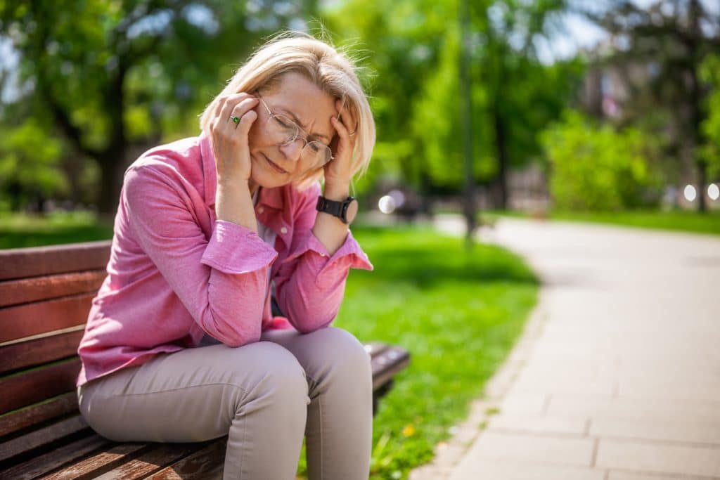 A woman sitting on a bench outside, holding her head in pain.