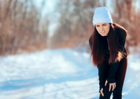 A woman out in the snow holding her knee in pain due to cold weather aches.